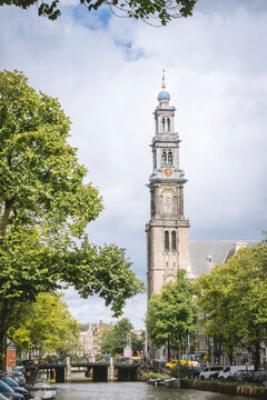 Tall clock tower in a European city with trees and a canal under a cloudy sky. Amsterdam,. Netherlands