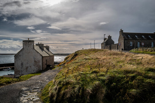 Coastal village with stone houses, cloudy sky, and a grassy path leading to the water's edge. Keiss harbour, Scotland
