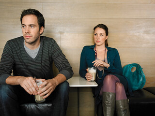 A man and woman sit separately in a cafe holding drinks, looking away with neutral expressions. Germany
