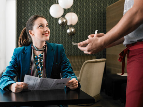 Woman in a teal suit sits at a table looking at a server holding notepad and pen. Germany