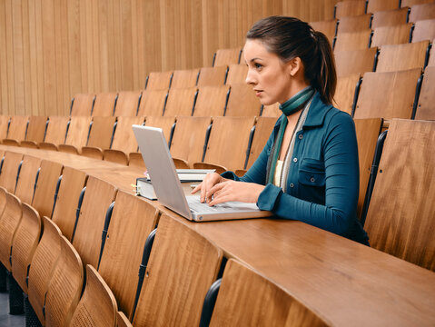 Woman using laptop in an empty lecture hall with wooden seats and books beside her. Germany