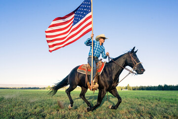A cowboy riding a horse holds an American flag in a green field under a clear blue sky. Oregon, USA