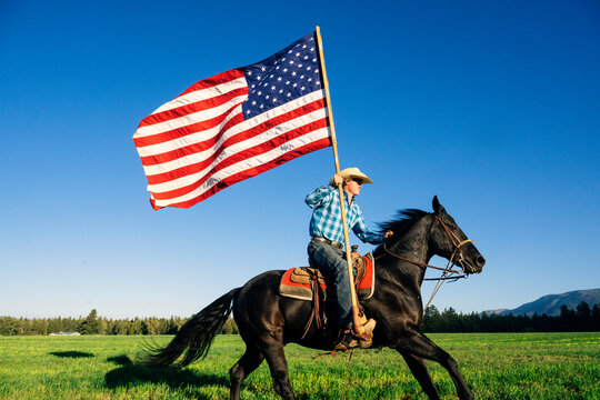 Cowboy on horseback carrying an American flag across a green field under a clear blue sky. Oregon, USA