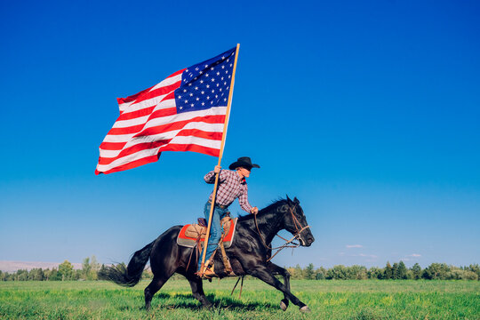 Cowboy on horseback waving large American flag in open green field under clear blue sky. Oregon, USA