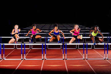 Five women competing in a hurdles race on a track at night. WA, USA