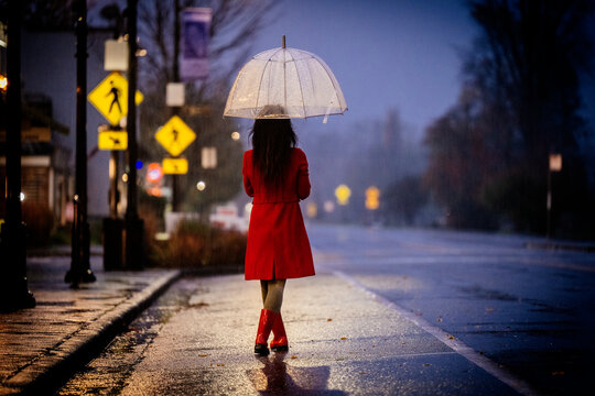 Woman in red coat and boots walks with umbrella on rainy street at dusk. WA, USA
