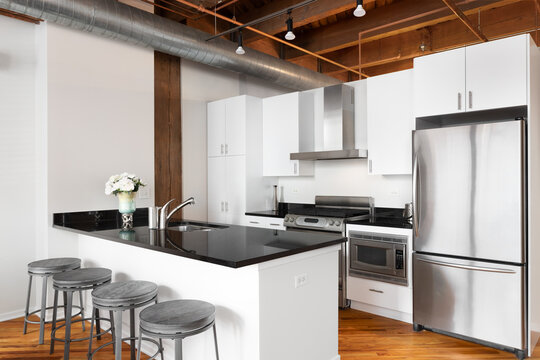 A loft white kitchen with exposed wood and ductwork ceiling, black granite countertops, bar stools sitting at the island, and stainless steel appliances. No brands or labels.