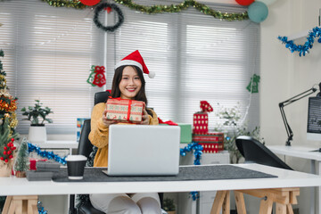 Young woman wearing santa hat holding wrapped gift smiling during festive office video call