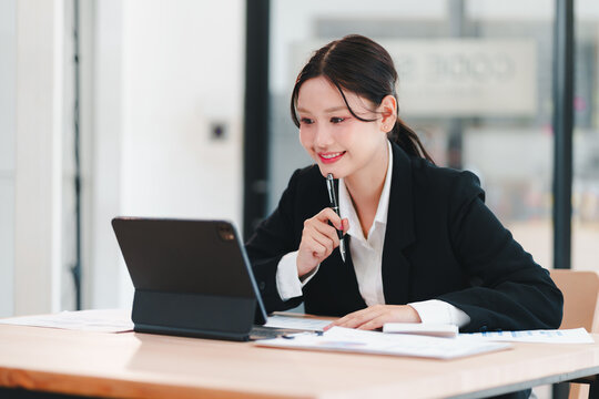 Young businesswoman in suit smiling while working on tablet and taking notes in modern office - Powered by Adobe
