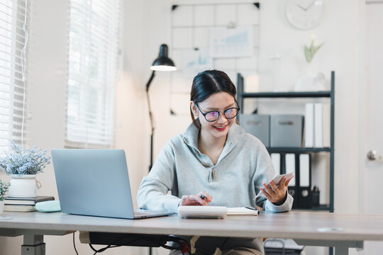 Young woman working at home desk smiling while using smartphone and laptop in bright home office
