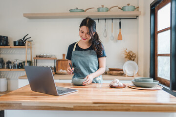 Young woman cooking in bright kitchen smiling while following online recipe on laptop