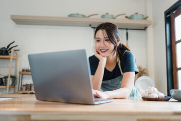 Young woman in apron smiling while using laptop in kitchen, bright natural light, casual home cooking scene