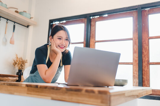 Young woman in apron smiling at laptop on kitchen island, warm morning light and cozy cooking scene - Powered by Adobe