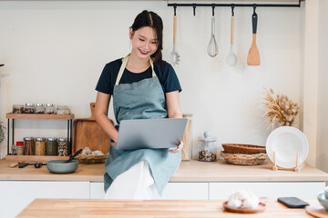 Young woman in kitchen apron using laptop while smiling, cozy home cooking scene with utensils and jars