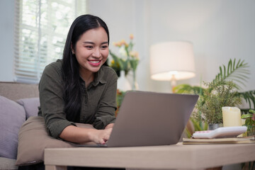 Young woman smiling while working on laptop at cozy living room table with plants and lamp