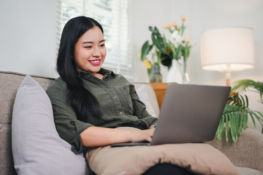 Young woman smiling while using laptop on sofa in cozy living room with plants and lamp