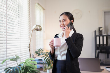 Young woman businessperson talking on phone while holding mug, smiling by sunlit office window