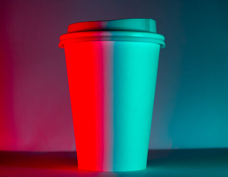 Neon lit takeaway coffee cup on dark background with red and cyan lighting. Minimalist cyberpunk style still life with strong contrast and modern aesthetic.