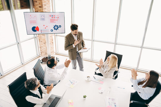 Team members applaud a colleague in a modern office meeting with charts and figures displayed on a screen.