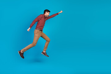 Obraz premium Young handsome man in casual shirt and tan pants leaps in mid air against blue background