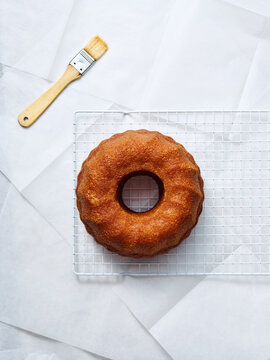 Freshly baked ring cake cools on a white wire rack, next to a basting brush. The cake sits against a backdrop of white parchment paper in a bright setting