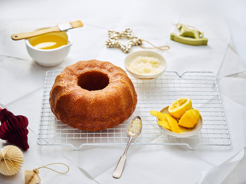 A ring cake rests on a wire rack with lemons, sugar, glaze, and ornaments around it. It looks like the cake is being prepared for decoration with a lemon glaze