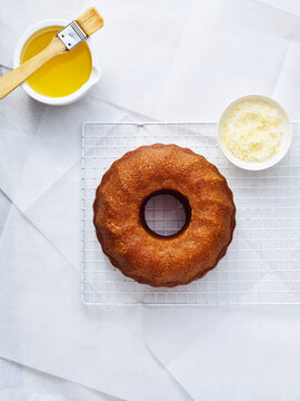 Ring cake rests on a cooling rack. Melted butter with a brush and shredded coconut are nearby, ready for preparation. Light reflects off the white surface
