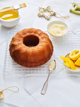 A golden bundt cake is cooling on a rack. Lemon glaze and lemon wedges surround it, and Christmas ornaments add a festive touch. Baking tools nearby suggest the cake was freshly prepared