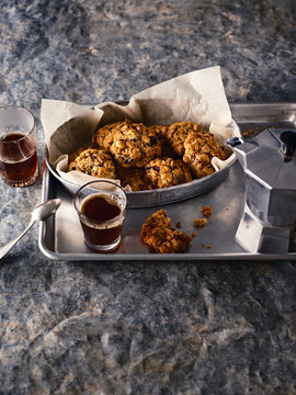 Fresh baked oatmeal cookies are presented on a metal tray with baking parchment. Also present are glasses of espresso and a moka pot against a textured, grey backdrop