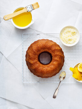 A freshly baked lemon bundt cake rests on a wire cooling rack. Bowls of melted glaze, citrus peel, and a pastry brush are positioned nearby. A spoon has a bit of glaze on it