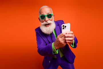 Energetic elderly man posing for a playful selfie in a purple suit and green sunglasses against an orange backdrop