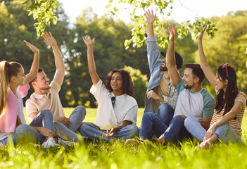Group of friends having meeting in summer park. Several happy young diverse people sitting on green grass, having fun, discussing sustainable living ideas, and raising hands up to vote or volunteer