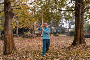 Man doing martial arts exercises with a wooden staff among fallen leaves in a park