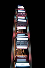 Close-up view from beneath the cabins of a Ferris wheel glowing with white and pink lights at night. The image focuses on the geometric lines and modern design against a pitch-black sky.