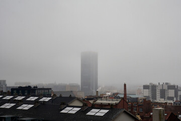 A dense morning fog blankets the rooftops of Brussels, Belgium, partially hiding a tall building in the background. The misty atmosphere creates a moody and mysterious cityscape