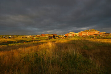 Golden rural landscape on Solovetsky Island with wooden houses, fields and dramatic storm clouds at sunset.