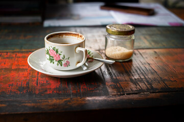 Perfect cup of black coffee served with a spoon and bottle of sugar on a simple white plate at red wooden table in a cafeteria. 