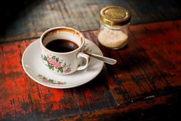 Perfect cup of black coffee served with a spoon and bottle of sugar on a simple white plate at red wooden table in a cafeteria. 