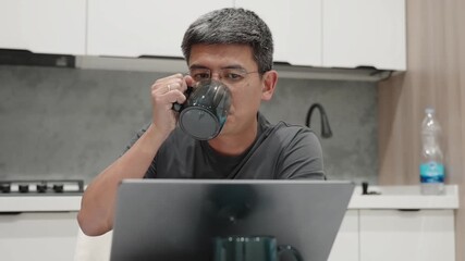 Asian man taking sip from dark glass mug while working on his laptop in  clean modern kitchen. The photo represents remote work habits, routine productivity, and a relaxed home atmosphere.