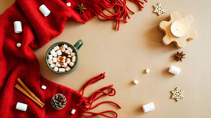 Overhead shot of a mug with marshmallows, a red scarf, and winter decorations on a beige surface