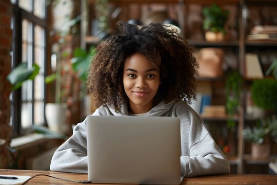 A young woman with curly hair sits at a desk in a warm, inviting space filled with greenery. She smiles confidently at her laptop while enjoying her work or leisure time