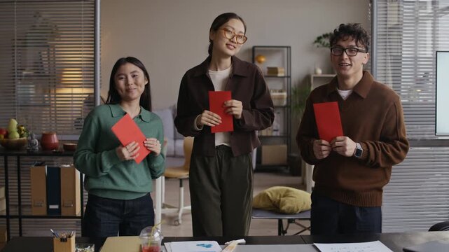 Medium long portrait of three Asian young colleagues with red envelopes looking at camera while celebrating Tet in modern office