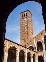 A low-angle shot captures the medieval red brick bell tower of the Basilica of Sant'Ambrogio in Milan, framed by a stone arch in the foreground.