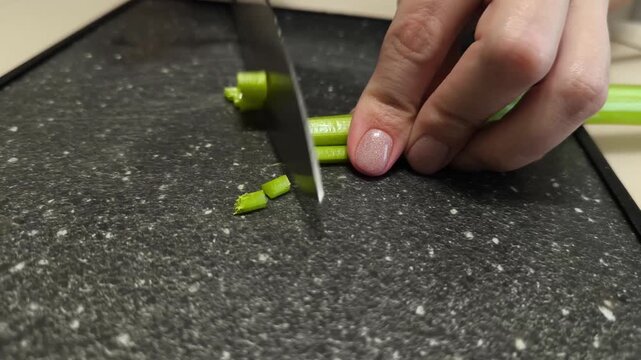 A woman's hand slices a celery stalk with a sharp knife on a dark cutting board. A close-up of the healthy cooking process.
