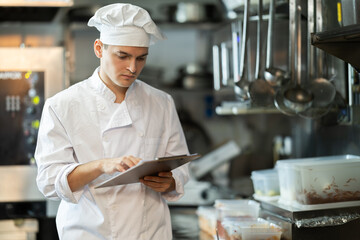 Young male chef in uniform with clipboard conducting inspection in restaurant kitchen