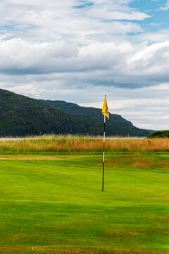 Yellow flag on green golf hole amid green course and hills under cloudy sky