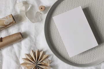 Christmas gift wrapping flat lay with blank card, craft paper ornaments, ribbon. White table cloth. Scandinavian holiday packaging, DIY craft stationery mockup, eco friendly festive styling, top view.