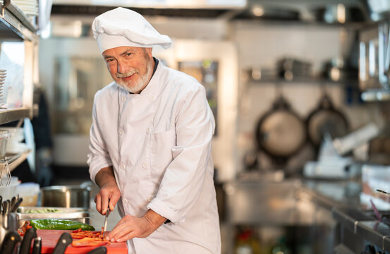 Senior man cook in kitchen finely slices food and prepares ingredients for cooking. Kitchen worker in white uniform performs tasks of chef and auxiliary functions - Powered by Adobe