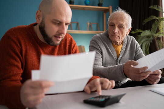 Caucasian middle-aged son helping old father counting expenses and pay bills at home.