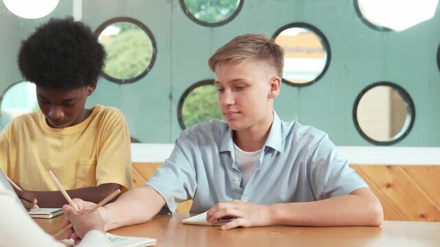 Young smart teenager writing and talking about topic at classroom. Diverse high school student taking a note in other notebook while sitting with multicultural friends. Creative education. Edification